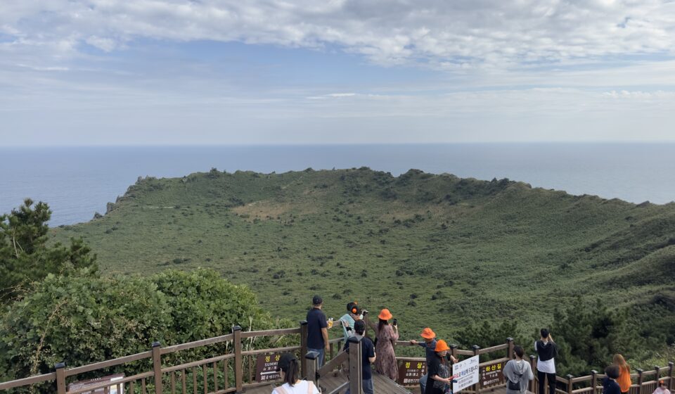 Panoramic view from the summit of 성산일출봉 in Jeju Island, South Korea, overlooking the coastline and surrounding sea.